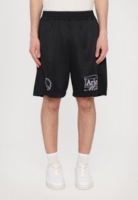 Shorts en maille noirs avec une taille élastique, ornés de logos brodés sur la cuisse gauche et de l'autre côté. Portés avec des baskets blanches.