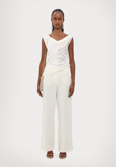 White draped top with a rounded neckline, paired with off-white wide-leg trousers. Model holds a metallic bag, wearing open-toed sandals.