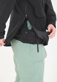 Musta vedenpitävä takki, jossa on vetoketjclosure ja taskuyksityiskohtia. Yhdistetty vaaleanvihreisiin housuihin, jotka ovat sileäpintaiset ja joissa on sivutasku.