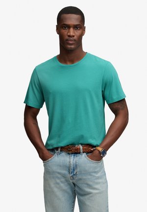 Uomo con t-shirt verde acqua a maniche corte, jeans azzurri chiari con cintura intrecciata marrone e orologio, in piedi con le mani in tasca su sfondo bianco.