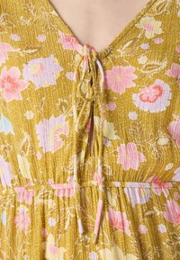 Robe fleurie avec une base jaune moutarde ornée de fleurs roses et bleues, un décolleté en V et un détail à cordon au niveau du col. Tissu texturé.