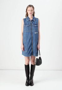 Denim bez piedurknēm kleita ar priekšējo rāvējslēdzēju, divām krūšu kabatām un izliekto apakšu. Apvienota ar melniem augstiem zābakiem līdz ceļgaliem un mazu melnu summiņu.
