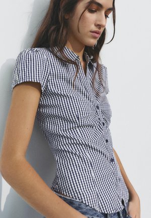 Žena opretá o stenu, oblečená v čierno-bielej kockovanej krátkoruká blúzka a modré džínsy, pozerá dole s uvoľneným výrazom.