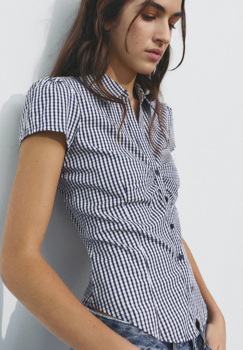 Femme appuyée contre un mur, portant une chemise à carreaux noir et blanc à manches courtes et un jean bleu, regardant vers le bas avec une expression détendue.