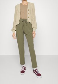 Beżowy sweter dzianinowy z guzikami, brązowy t-shirt, oliwkowe spodnie z sznurkiem, oraz bordowe trampki z białą podeszwą.