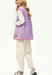 Kobieta o długich blond włosach, ubrana w pikowaną lawendową kamizelkę z pomarańczowymi obszyciami, luźne kremowe spodnie, biały długi rękaw i beżowe trampki.