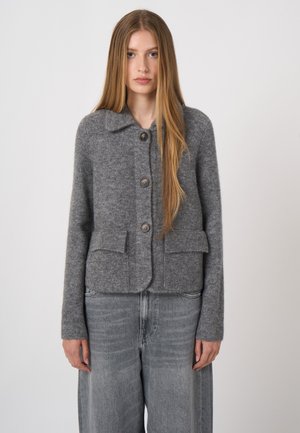 Junge Frau mit langen blonden Haaren trägt eine graue wollene Jacke mit Knöpfen und lockere graue Jeans vor einem schlichten hellen Hintergrund.
