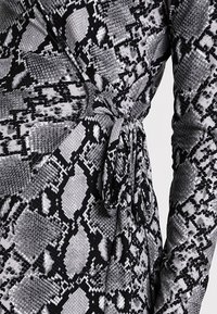 Zbliżenie na sukienkę w wężowy wzór w odcieniach czerni i szarości, przewiązaną w talii, z długimi rękawami.