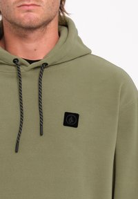 Olijfgroen fleece hoodie met zwarte trekkoorden, met een vierkante zwarte logopatch op de linkerborst en een zachte textuur.