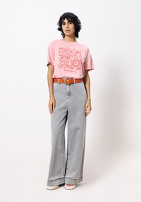 Roze grafische t-shirt met korte mouwen, voorzien van een rode opdruk. Lichtgrijze wijde spijkerbroek met rafelige zoom. Model draagt een oranje riem.