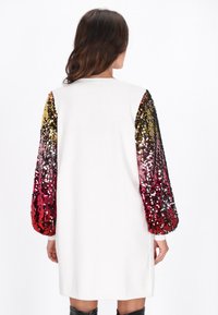 Sukienka z long-sleeve w białym materiale, z rękawami zdobionymi cekinami w przejściach od złota do czerwieni. Okrągły dekolt i luźny krój.