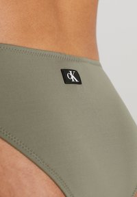 Olivgrön bikiniunderdel i slät tyg med en svart CK-logotypapplikation. Har rena sömmar och en låg midja.
