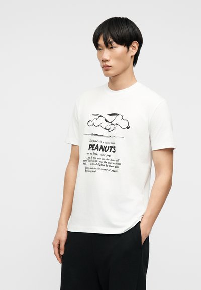 Νεαρός με μαύρα μαλλιά που φοράει λευκό μπλουζάκι με μαύρο γραμμικό σχέδιο του Snoopy από τα Peanuts και κείμενο κάτω από την εικόνα.