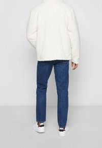 White fleece jacket with a relaxed fit, blue denim jeans, and white sneakers featuring black accents. Textured surface and round collar.