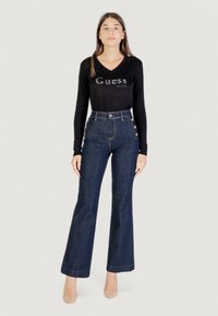 Maglia nera a maniche lunghe con logo "Guess" in paillettes; jeans flare in denim scuro a vita alta con bottoni laterali e texture liscia.