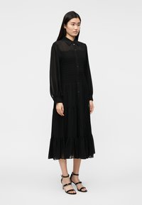 Čierne šaty s gombíkovým zapínaním vpredu, dlhými rukávmi a vrstvovým, volánkovým lemom. Vyrobené z ľahkého materiálu so smockovaným živôtikom.