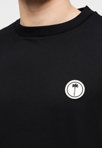 Μαύρη μπλούζα με στρογγυλή λευκή επιγραφή με μαύρη φοινικιά και κείμενο στην αριστερή πλευρά του στήθους.