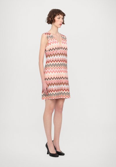 Patterned sleeveless dress featuring a V-neckline, zigzag design in shades of pink, brown, and white, paired with black high heels.