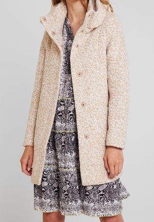Femme portant un manteau beige texturé à boutons sur une robe à motifs noir et blanc avec des touches jaunes, debout devant un fond uni.
