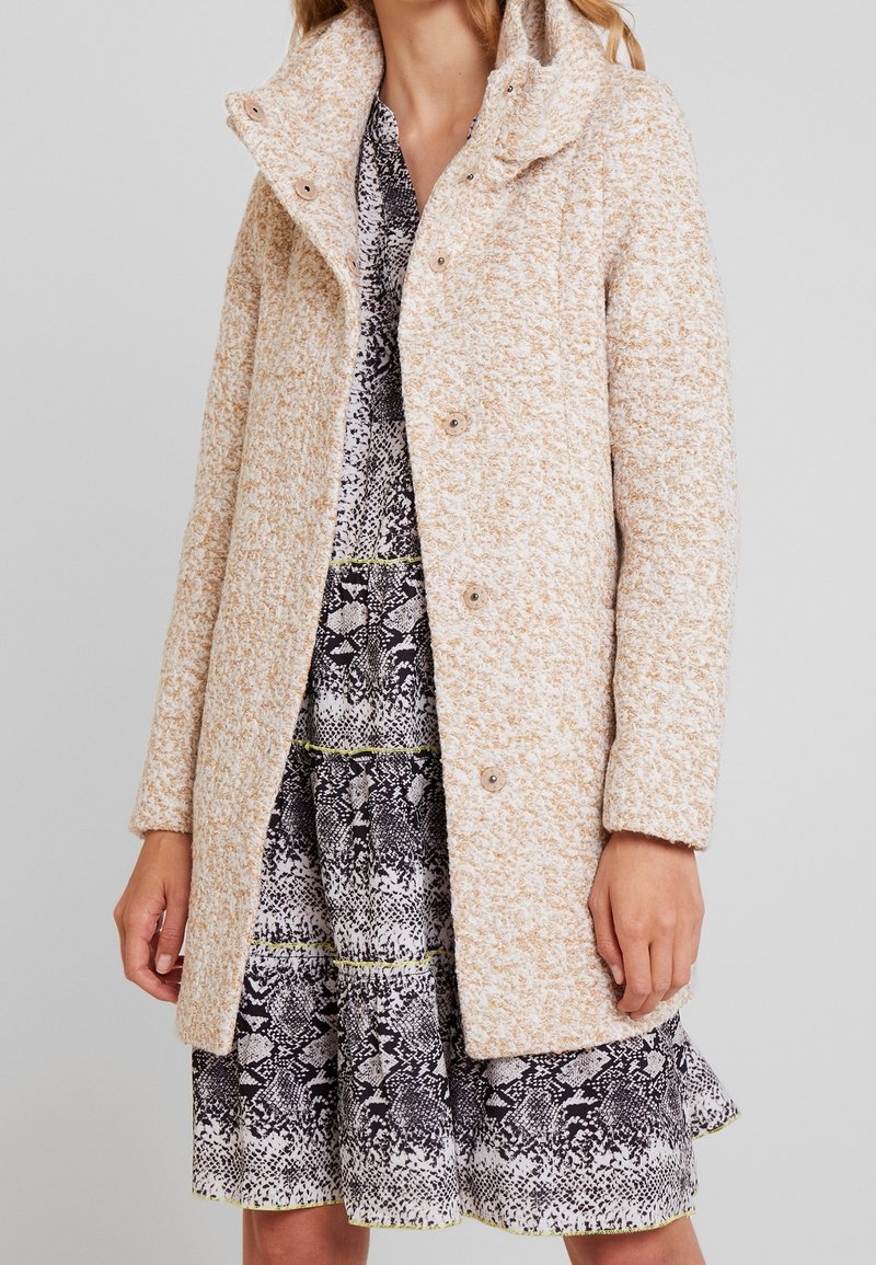 Femme portant un manteau beige texturé à boutons sur une robe à motifs noir et blanc avec des touches jaunes, debout devant un fond uni.