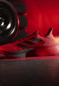 Basket de sport rouge et noire avec trois bandes noires, placée sur une surface à côté d'une plaque de poids noire sur fond rouge.