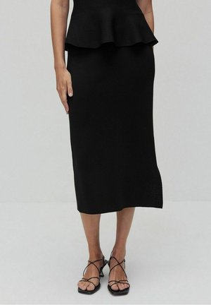 Femme portant une jupe crayon noire longueur genou et des sandales à talons noires avec brides, debout devant un fond clair uni.