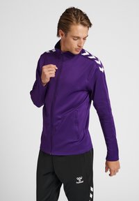 Giacca sportiva viola con colletto alto e zip completa; presenta accenti bianchi a zigzag sulle spalle e tasche laterali.