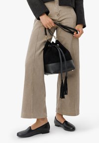 Sac à seau en suède noir avec une base en cuir texturé, orné de franges et de pièces métalliques dorées, tenu par une personne portant un pantalon beige et des chaussures noires.