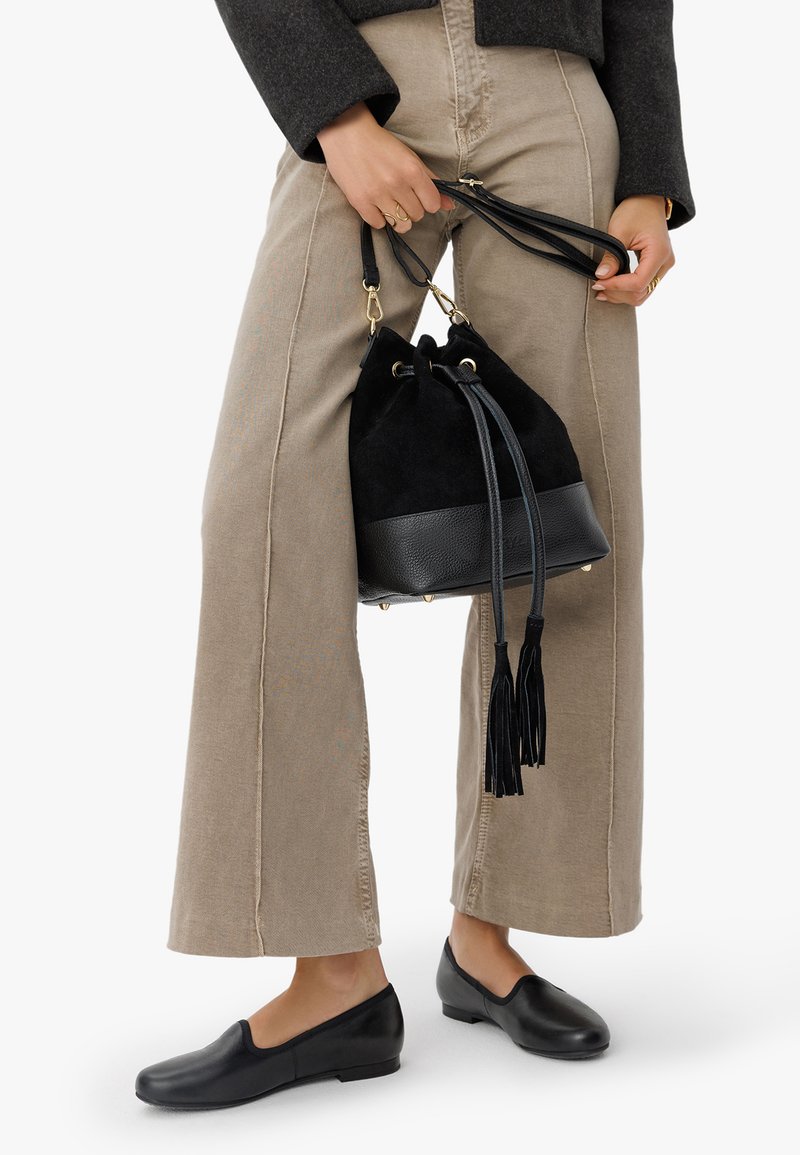 Sac à seau en suède noir avec une base en cuir texturé, orné de franges et de pièces métalliques dorées, tenu par une personne portant un pantalon beige et des chaussures noires.