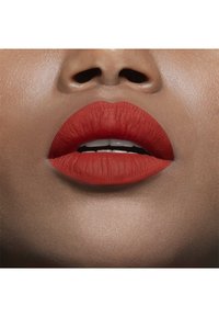 Rouge à lèvres mat rouge appliqué sur des lèvres pulpeuses, avec une texture lisse. Le teint est chaud et uniforme, avec un léger éclat à la surface.