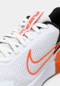 Baskets blanches de sport avec une tige en mesh, un swoosh Nike orange, des accents gris et une semelle texturée. Présente des lacets blancs et une languette avec un onglet rose.