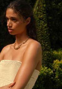 Collier en chaîne en or et en argent avec des maillons allongés porté avec un haut sans manches à texture légère, sur fond vert en plein air.