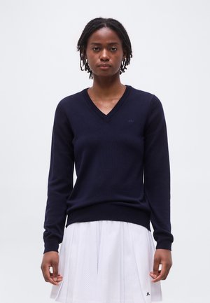 Kobieta z krótkimi dredami ubrana w granatowy sweter z dekoltem w serek i białą plisowaną spódnicę, stojąca na tle jasnego, jednolitego tła.