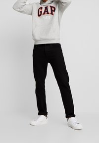 Sudadera con capucha gris con el logo "GAP PARIS" en rojo y azul, combinada con pantalones vaqueros negros y zapatillas blancas. La sudadera tiene un bolsillo tipo canguro.