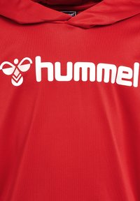 Czerwona bluza z kapturem wykonana z gładkiego materiału, z dużym białym logo "hummel" oraz charakterystyczną grafiką pszczoły z przodu.