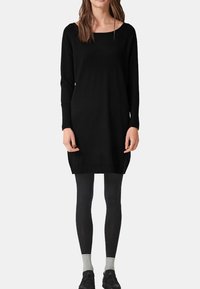 Donna che indossa un abito nero a maniche lunghe fino al ginocchio, leggings neri, calzini grigi e scarpe nere, in piedi contro uno sfondo bianco.
