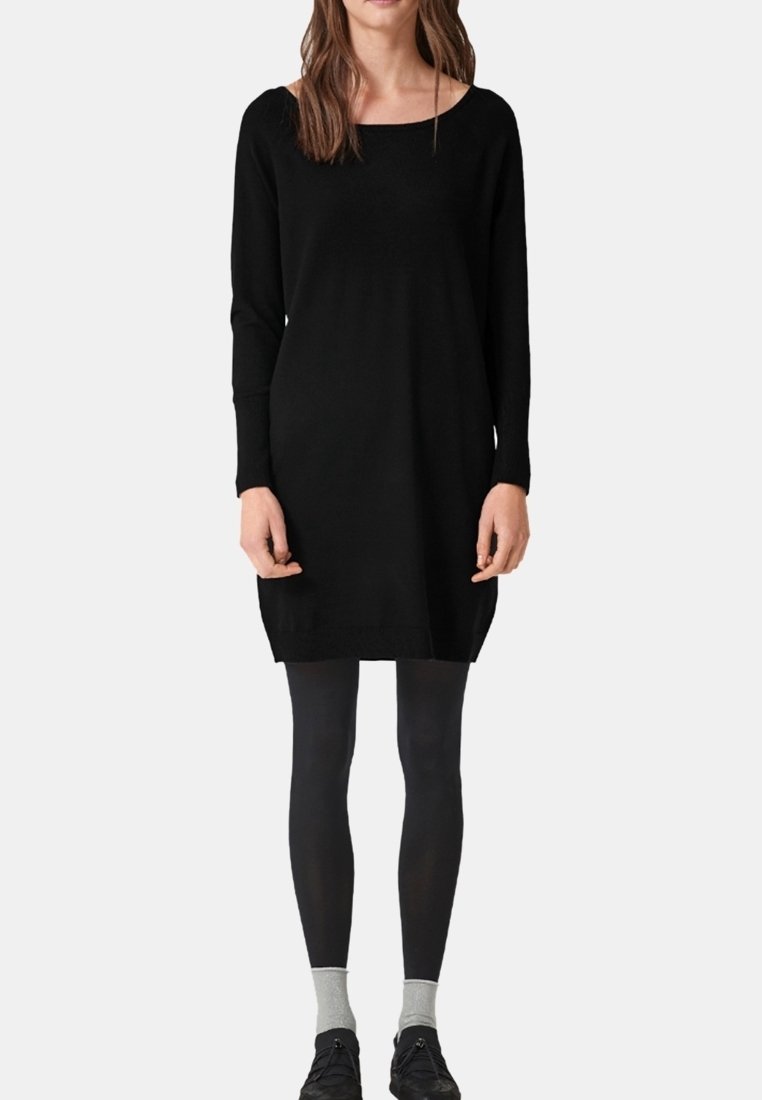 Donna che indossa un abito nero a maniche lunghe fino al ginocchio, leggings neri, calzini grigi e scarpe nere, in piedi contro uno sfondo bianco.