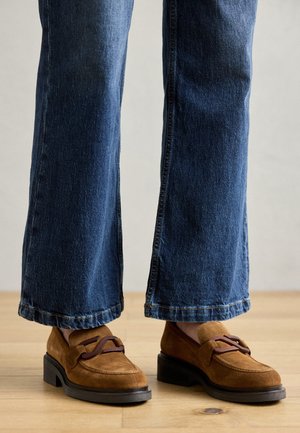 Mocasines de ante marrón con una tira decorativa, combinados con jeans de pierna ancha azules. Los jeans tienen un dobladillo deshilachado y están ligeramente desvaídos.