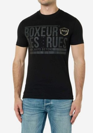 Mann trägt ein schwarzes eng anliegendes T-Shirt mit dem Schriftzug und Emblem „BOXEUR DES RUES“, kombiniert mit hellblauen Jeans, steht vor einem schlichten Hintergrund.