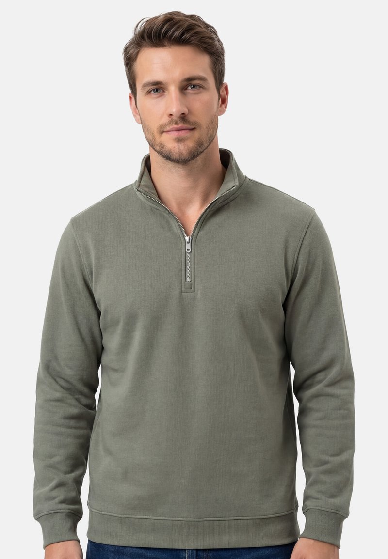 Homme portant un pull demi-fermeture éclair vert olive, debout devant un fond clair uni, face à l'avant avec une expression neutre.