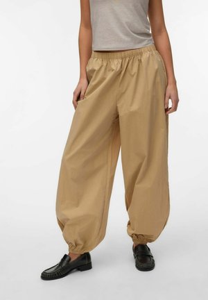 Femme portant un pantalon beige ample à taille élastique avec des chevilles reversées et des mocassins noirs, debout devant un fond blanc uni.