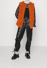 Veste sans manches orange duveteuse portée sur une chemise noire à manches longues en dentelle, associée à un pantalon cargo en cuir noir et des chaussures noires à lacets.