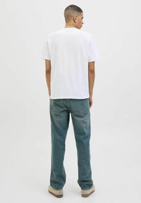 Hvid bomulds-t-shirt med korte ærmer, afslappet pasform. Båret med lyseblå denimjeans og beige sko, set fra ryggen. Ensfarvet baggrund.