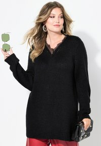 Schwarzer Strickpullover mit V-Ausschnitt und Spitzen-Details. Lange Ärmel, gerippte Bündchen und lockerer Schnitt. Getragen mit roten Hosen und grünen Sonnenbrillen.