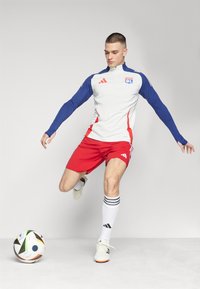 Muška sportska odjeća koja uključuje bijelu i plavu majicu dugih rukava s crvenim detaljima, uparena s crvenim šorcima i bijelim čarapama s crnim prugama, dok udara nogometnu loptu.