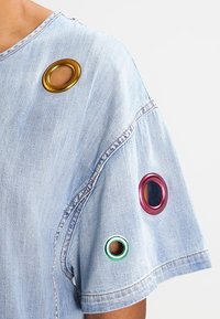 Ljusblå denimskjorta med ärm, prydd med tre metallringar i guld, rosa och grönt, som visar hud genom hålen.