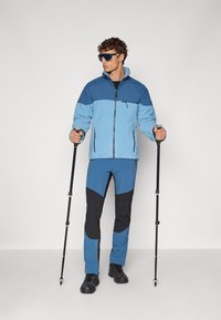 Giacca in pile di blu chiaro e scuro con chiusura a zip; abbinata a pantaloni blu e neri. Tiene racchette da trekking, indossa occhiali da sole.