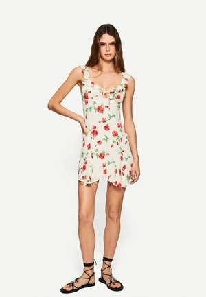 Robe fleurie avec des bretelles à volants et un détail en trou de clef. Tissu beige clair avec des roses rouges et des feuilles vertes. Ourlet court et évasé.