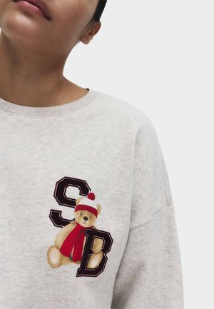 Sweatshirt gris clair présentant un ours portant un chapeau rouge et blanc, avec deux lettres bordeaux foncé "S" et "B" brodées à l'avant.