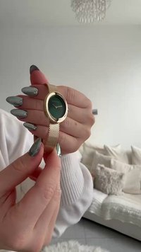 Hand mit langen, metallisch-silbernen Nägeln, die eine goldene Uhr mit Mesh-Armband und grünem Zifferblatt ohne sichtbare Zahlen hält.